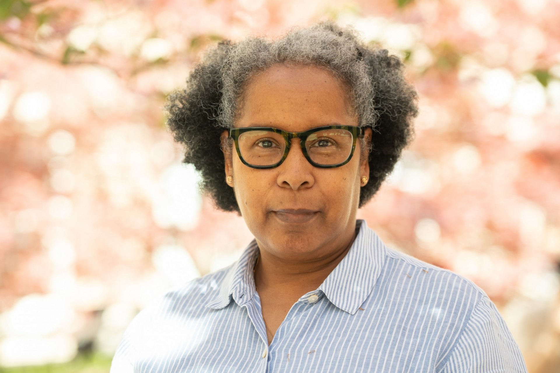 A headshot of a woman wearing glasses and a striped button-up, against a backdrop of cherry blossoms.