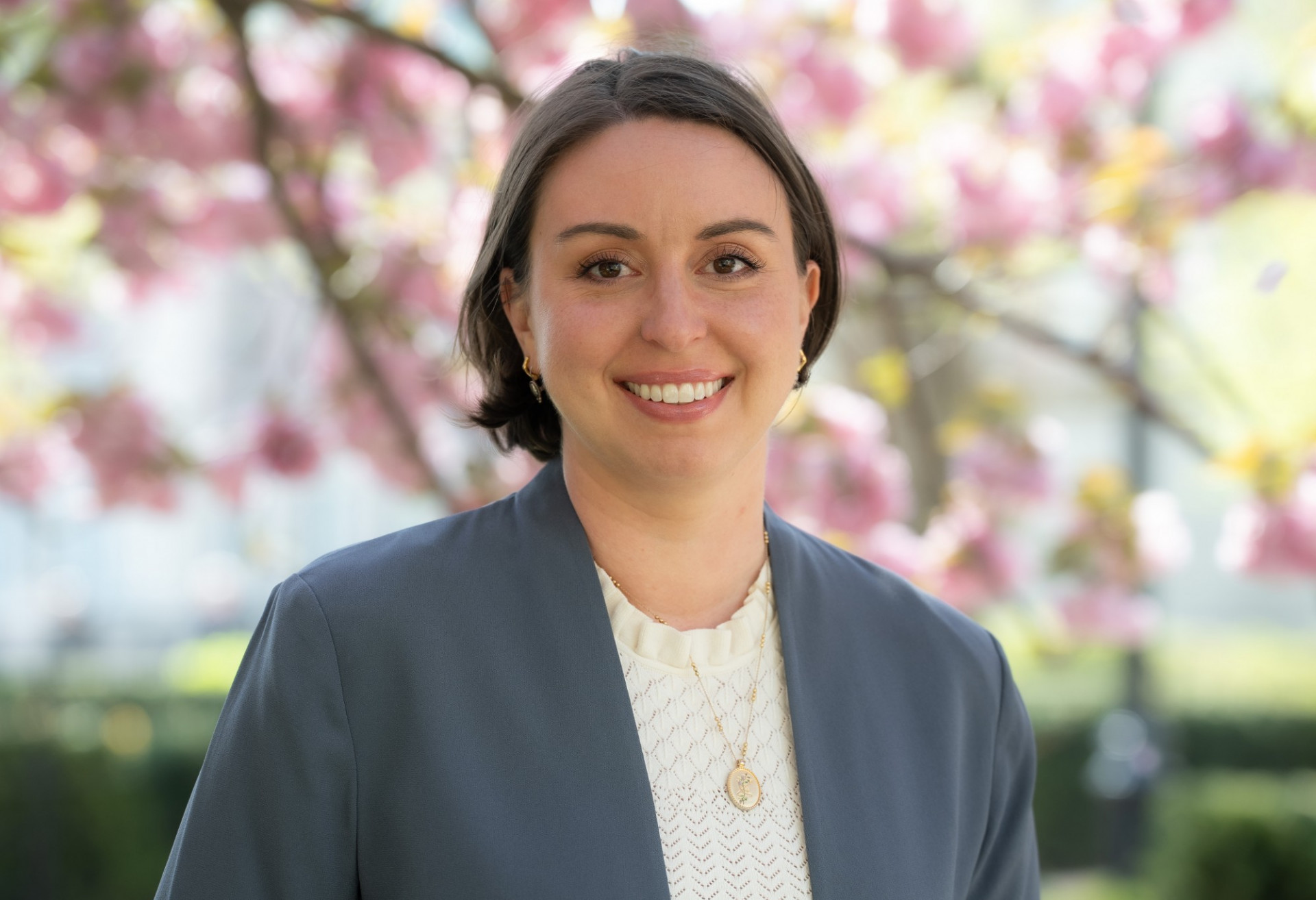 A woman with short brown hair in a gray blazer smiles against a cherry blossom tree.