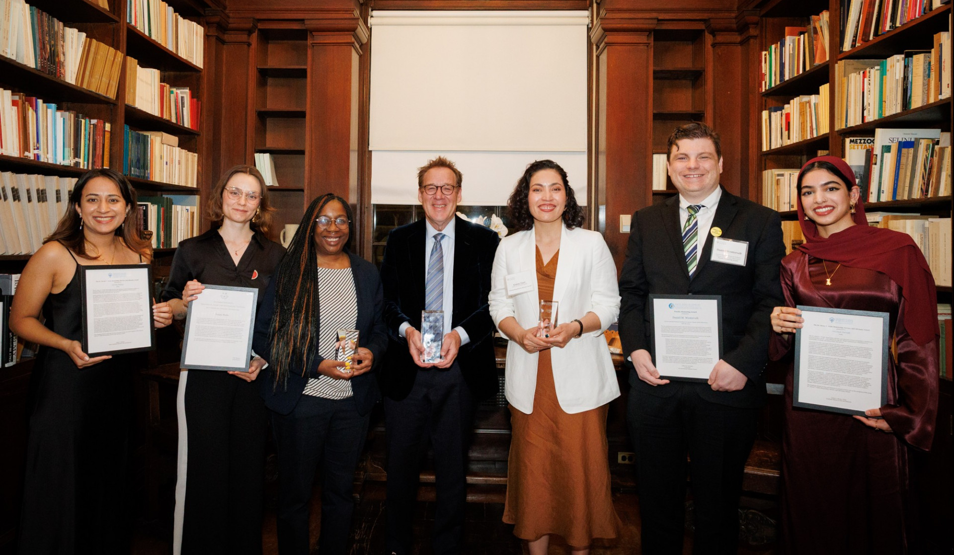 Seven people smiling in a library, holding awards and certificates. They stand between bookshelves filled with colorful books, conveying a celebratory mood.