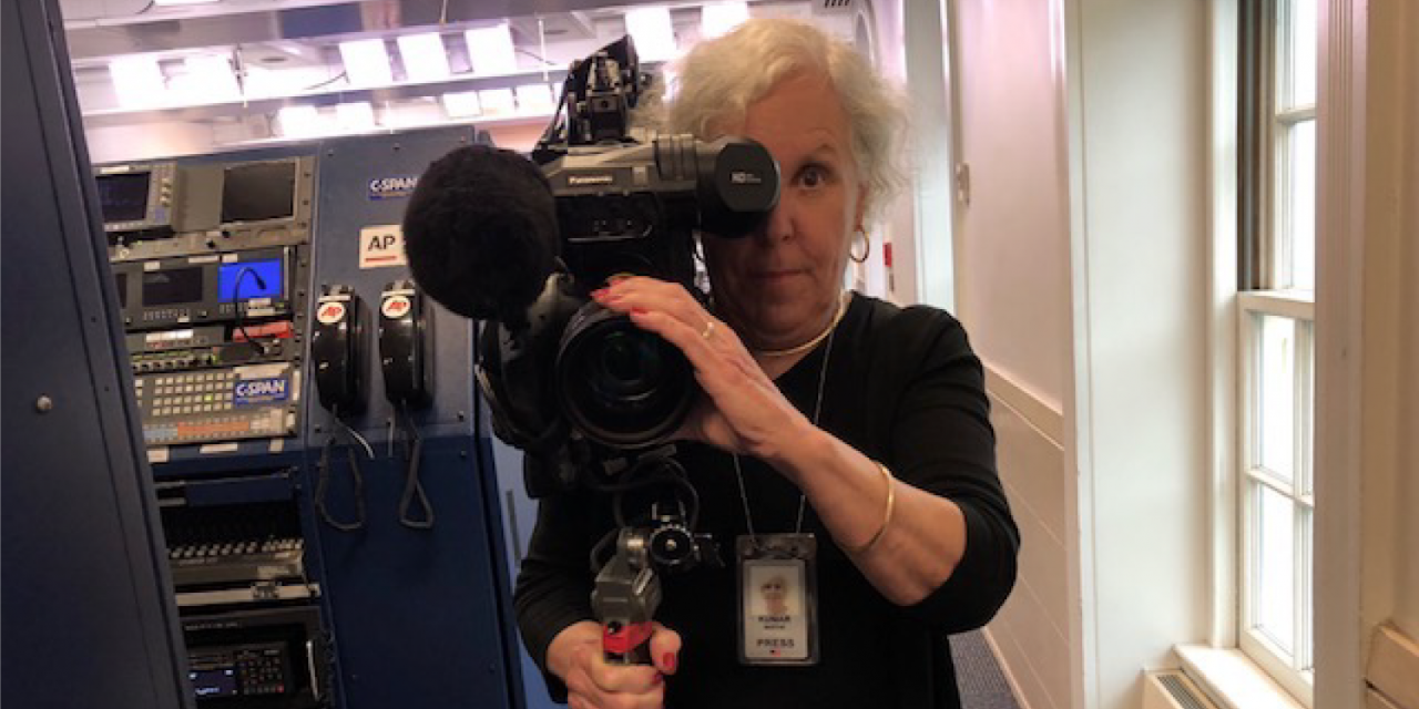 A woman with white hair looks at the camera through a video camera.