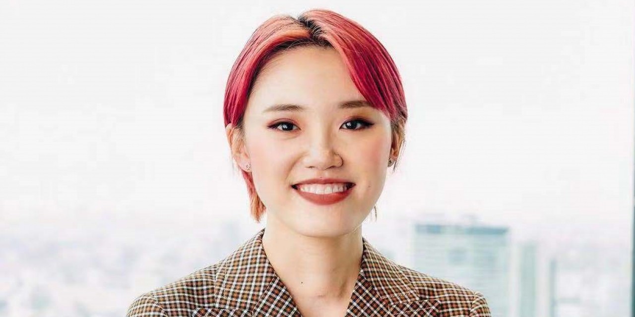 A woman with pink hair wears a checked blazer in front of a city backdrop.