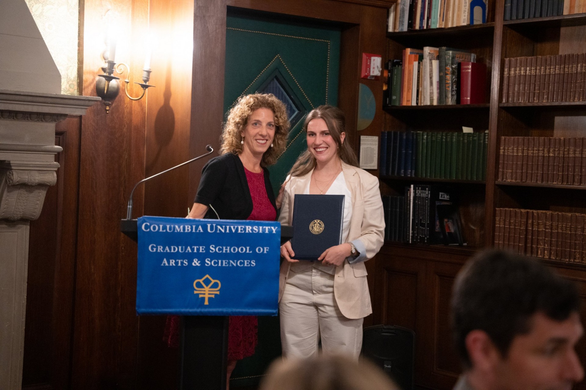 Two women at lectern, facing camera, one holding an award