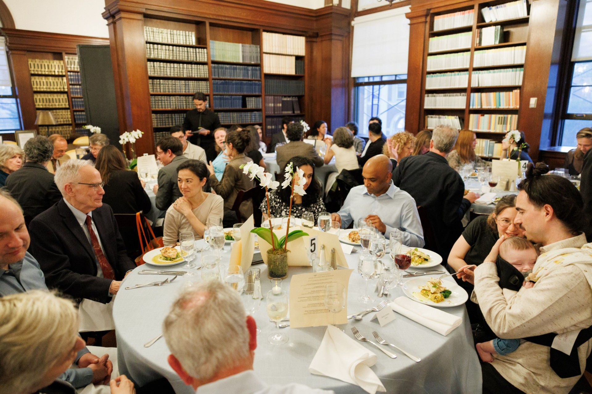 Students, alumni, and faculty enjoying the awards dinner in the library of Casa Italiana. Several tables with people seated all around, enjoying talking.