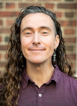 Fairskinned person with dark brown shoulder-length hair, with septum piercing and large jewel drop earrings, wearing purple button-down shirt with small white polkadots, closed-mouth smile