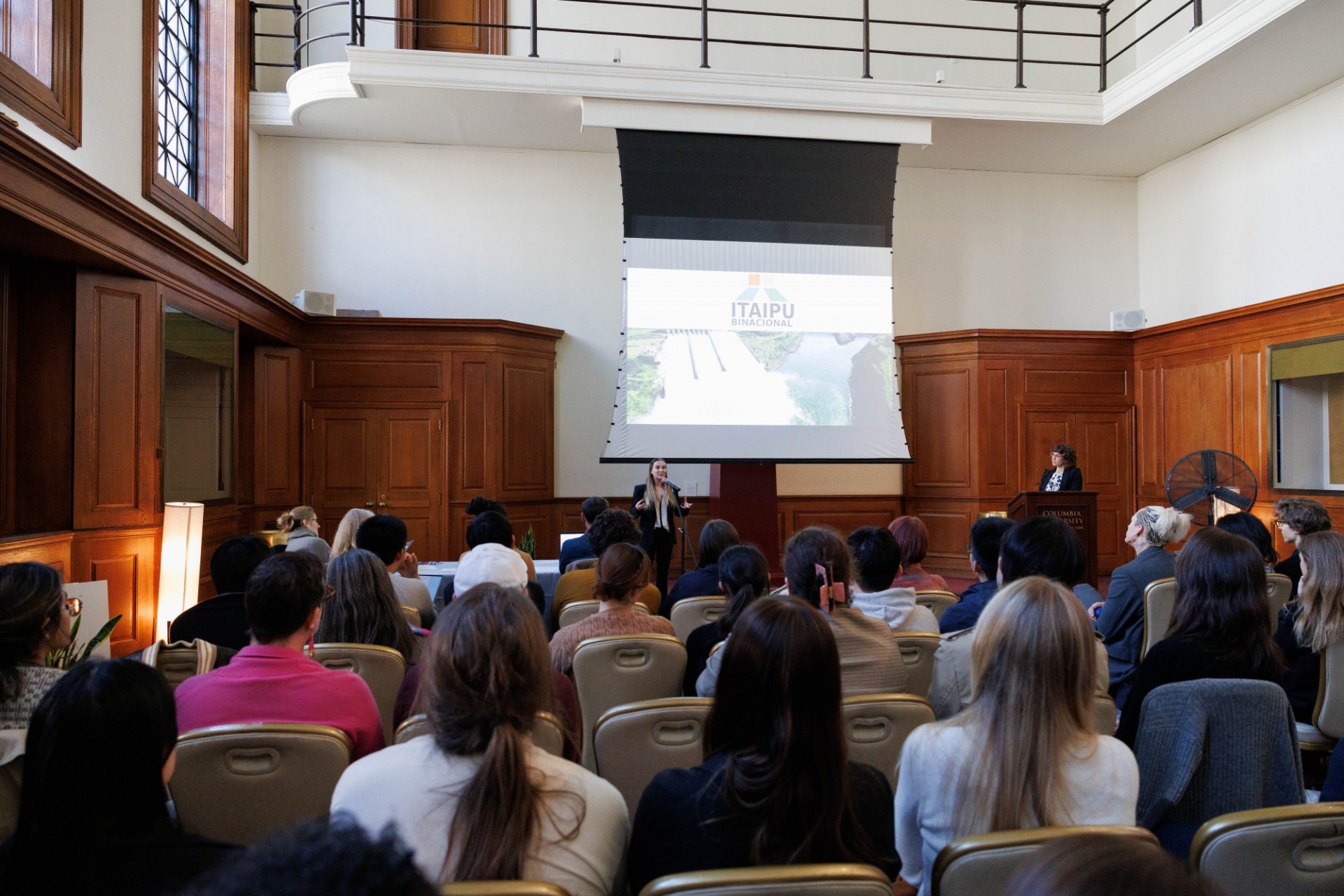 Agustina Carvallo (Quantitative Methods in the Social Sciences) gives a talk about the environmental impact of a dam in Paraguay wide shot from back of room full of people. Woman in front giving presentation.