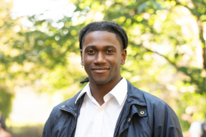 Head and shoulders of young man with dark skin, dark braided hair, white shirt and blue jacket smiling to camera