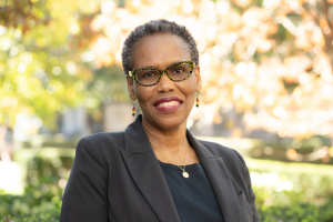 Head and shoulders of woman with dark skin, short dark hair, glasses and black shirt with dark suit jacket. She is smiling to camera. Campus greenery in background.