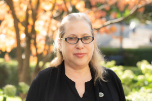 Head and shoulders of woman with glasses, light skin, dark shirt and and brown shoulder-length hair looking at camera