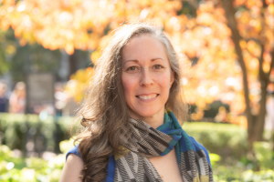 Head and shoulders of woman with long brown curly hair, wearing a scarf and smiling to camera.Campus greenery in background.