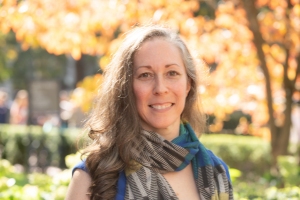 Head and shoulders of woman with long brown curly hair, wearing a scarf and smiling to camera.Campus greenery in background. 