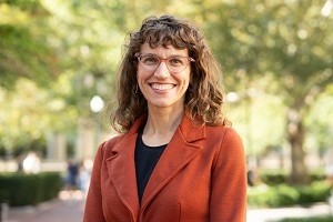 Head and shoulders of woman with curly brown shoulder-length hair with bangs, wearing glasses and smiling for the camera. Black shirt and orange suit jacket.