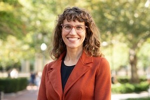 Head and shoulders of woman with curly brown shoulder-length hair with bangs, wearing glasses and smiling for the camera. Black shirt and orange suit jacket. 