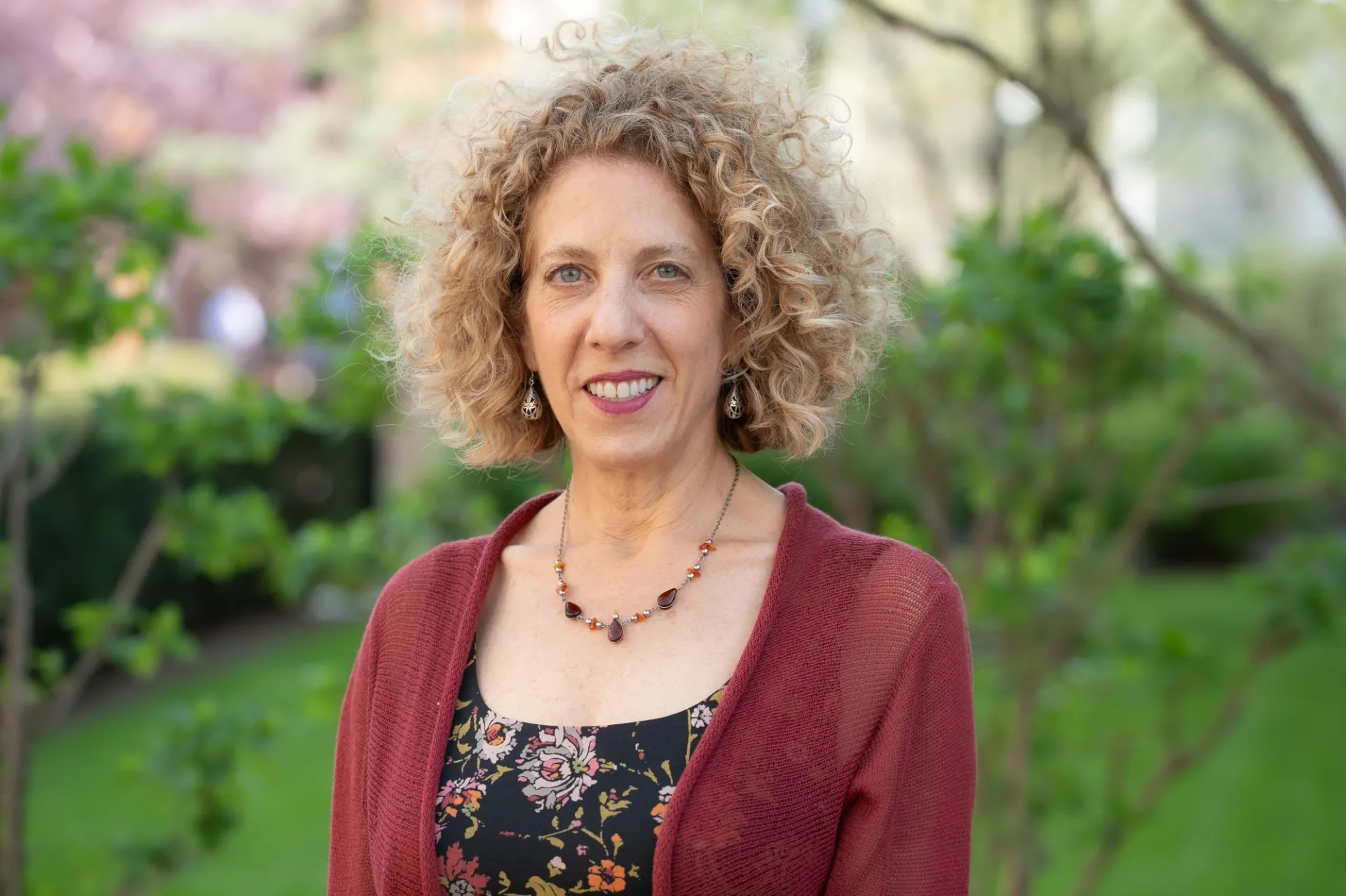 A woman with blond curly hair wearing a red sweater smiles against a backdrop of trees and grass. 