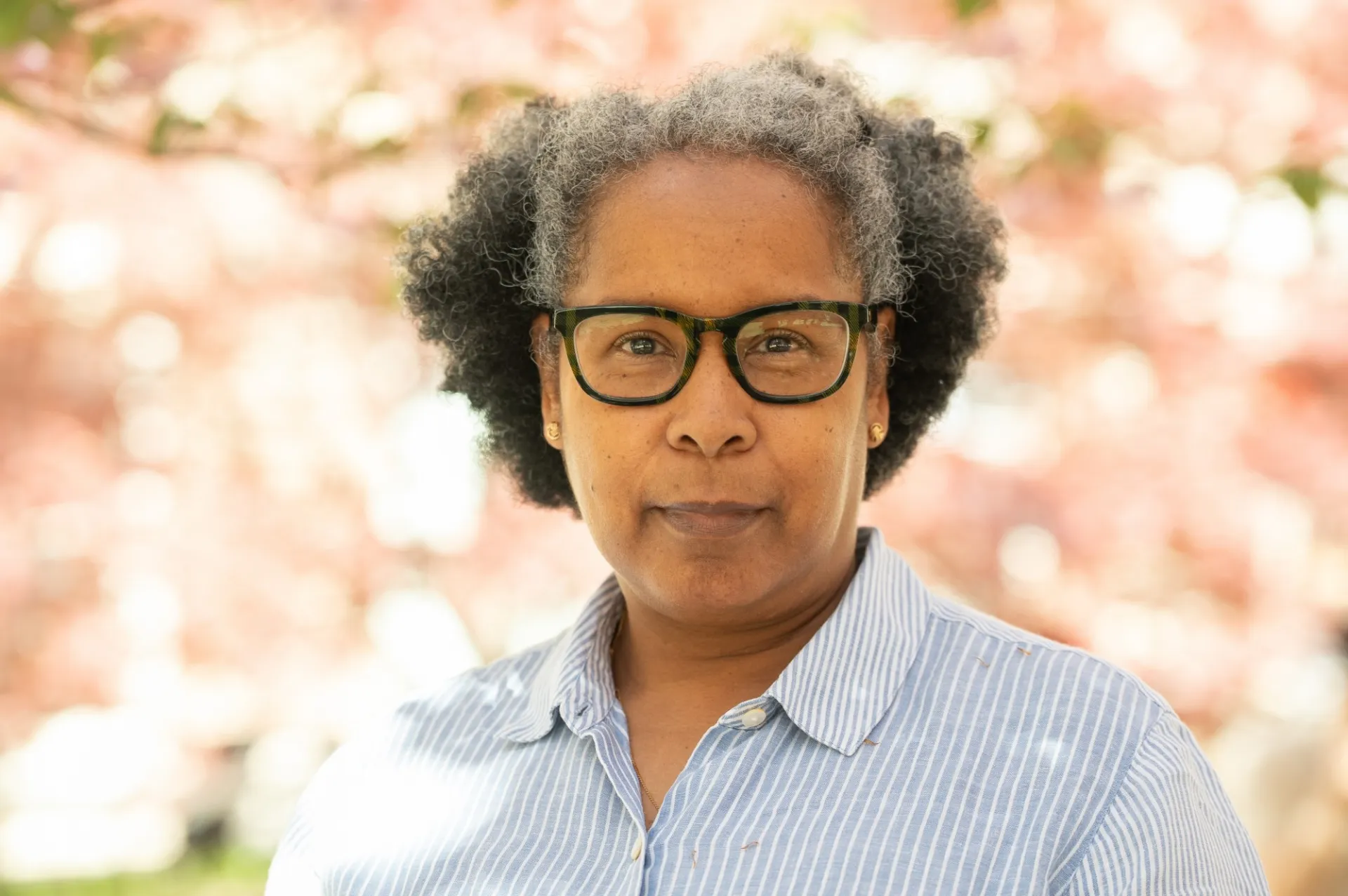 A headshot of a woman wearing glasses and a striped button-up, against a backdrop of cherry blossoms.