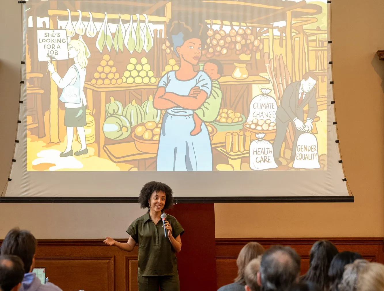 A woman speaking in front of a slide that shows an illustration of a woman and child at a market.