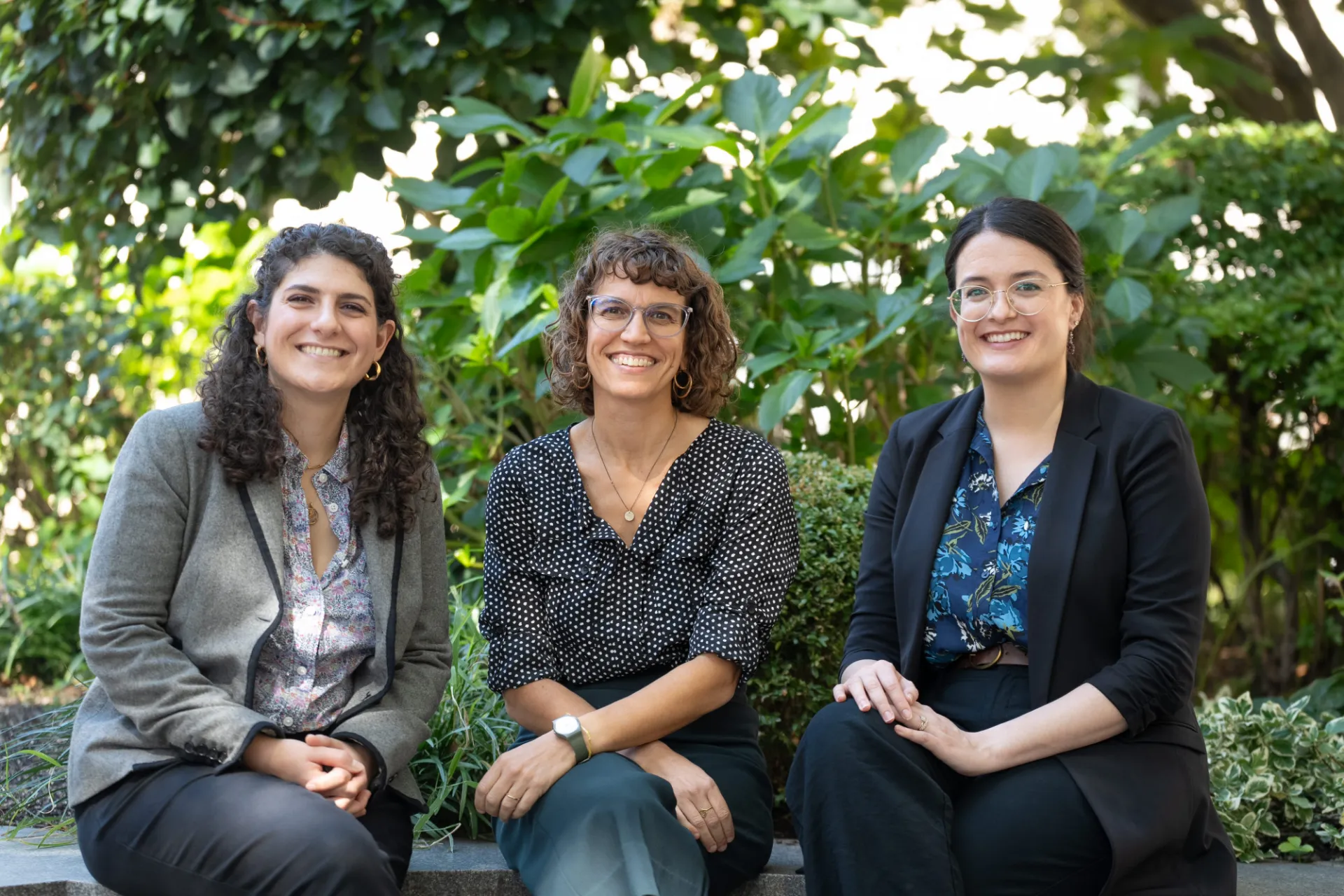 Three women are sitting on a stone bench in a lush garden, smiling confidently at the camera, dressed in professional attire.