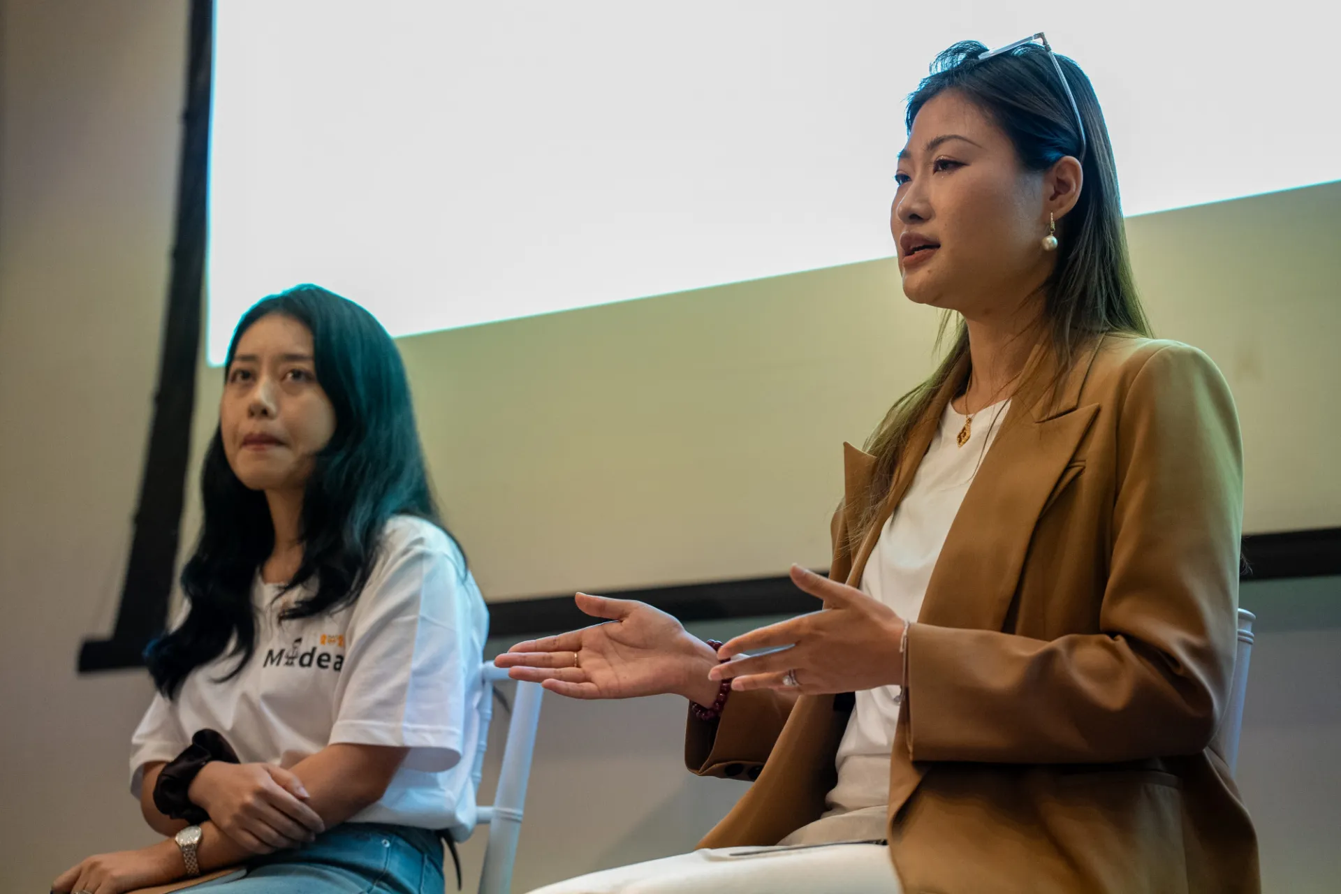 Two women are seated in front of a projector screen. The woman on the right, wearing a brown blazer, is speaking with expressive hand gestures. The woman on the left, in a white shirt, appears attentive. The setting suggests a formal discussion or presentation.