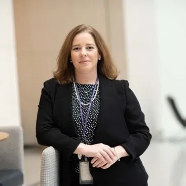 A woman with medium-length brown hear wearing a blazer, her hands clasped, leans on a shelf. A professional head shot.