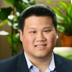 A man with short black hair smiles. He is wearing a black blazer. Plants are behind him, a sunny background.