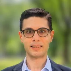 A man with glasses, short brown hair, wearing a suit, smiles for the camera.