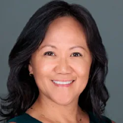 A woman with long brown hair smiles for a photo, a gray background. 
