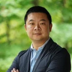 A man with short cropped hair wearing a blazer and blue shirt against a green backdrop. His arms are crossed.