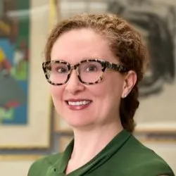 A woman with curly hair gathered in a bun and cat-eye thick glasses wearing a green shirt. She is smiling.