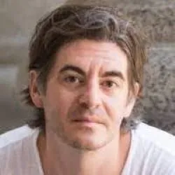 A man with tousled brown hair stares seriously into the camera. He is wearing a white shirt and in front of a stone background.