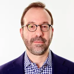 A man with round glasses and brown hair, with a light beard, wearing a blazer and a striped button-up.