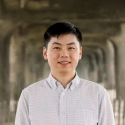 A young man with brown hair wearing a light-colored button-up smiles at the camera. 