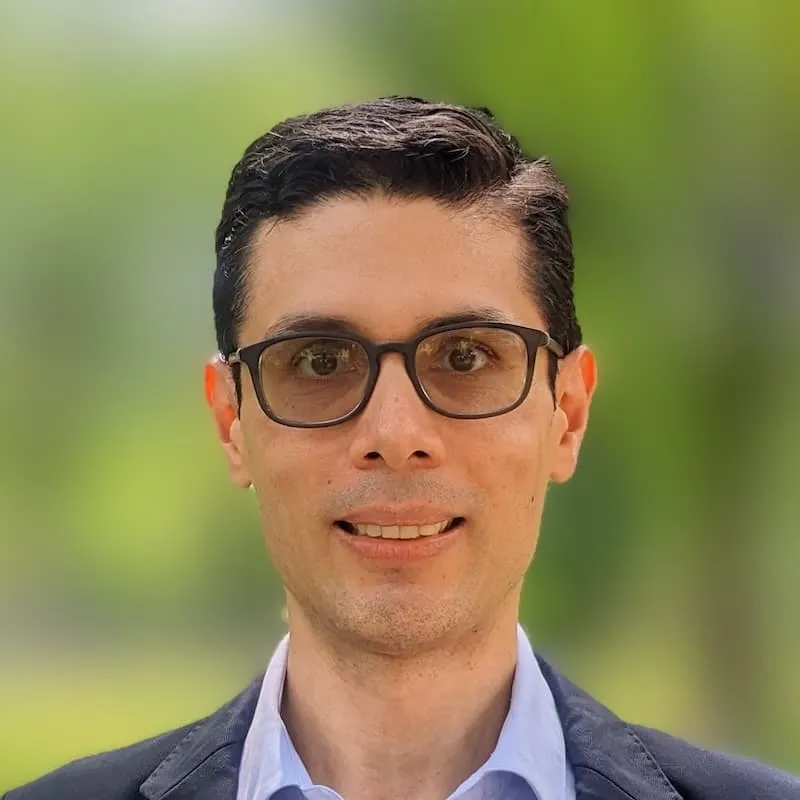 A man with brown hair and lightly tinted glasses wearing a blazer with a green backdrop.