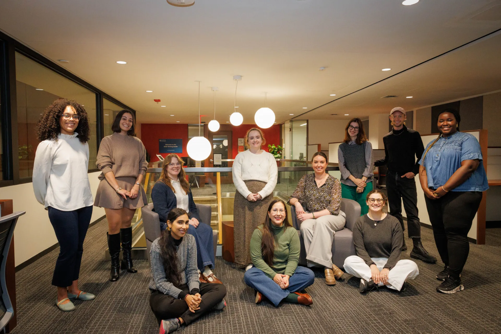 A diverse group of students and employees of the Writing Studio pose for a group shot in front of the stairwell, some are seated, some standing, all smiling for the camera.