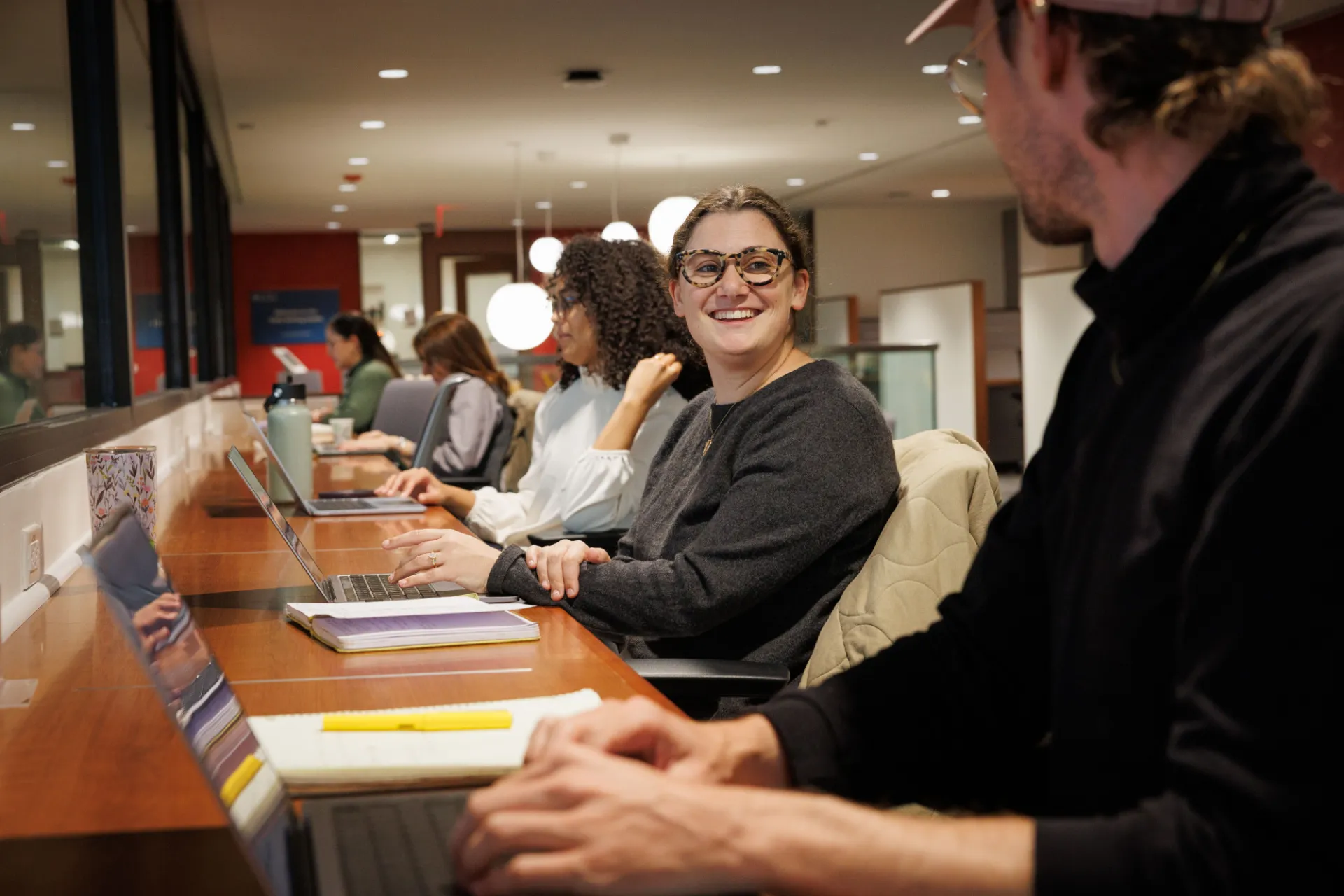 A woman wearing glasses sits at a long table with her laptop open in front of her. Beside her are other students writing on their laptops in a shared student space with bright lights and a window.