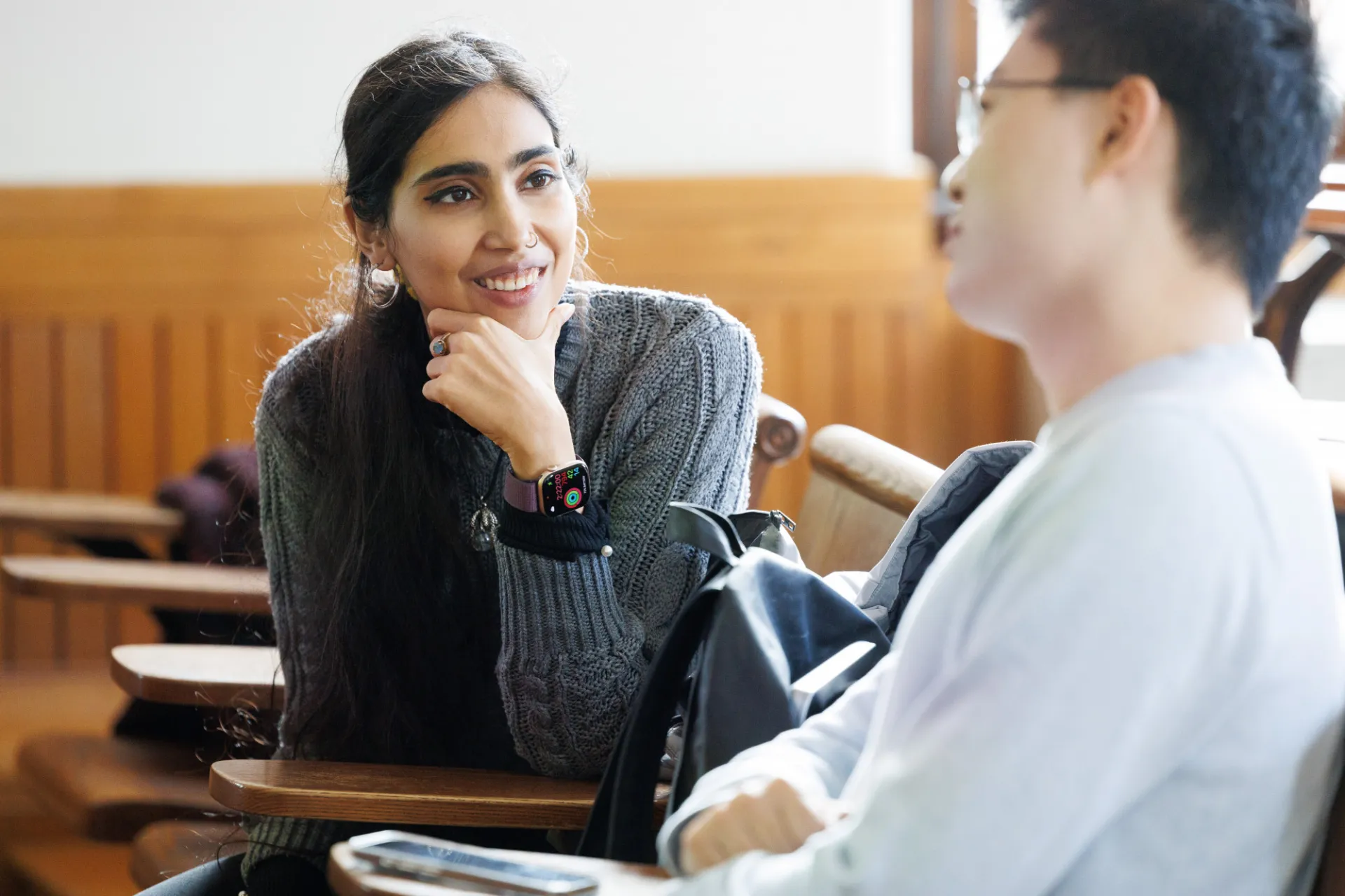 An instructor is seated at a desk in a classroom, beside her student. She is leaning with one hand propping up her chin, with a smile on her face as her student in the foreground speaks to her. There is a backpack on a chair between them.
