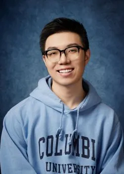 A smiling person wearing glasses and a light blue Columbia University hoodie poses against a soft, textured blue background, conveying a cheerful and academic tone.