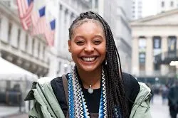Smiling woman with long braids stands outdoors in front of a building with columns. Wearing a patterned scarf and green jacket, conveying a joyful mood.
