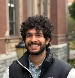 Smiling man with curly hair and beard in a black jacket, standing outside in front of a historic brick building. The mood is cheerful and relaxed.