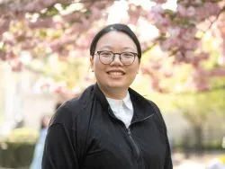 A person with glasses smiles while standing outdoors under blooming cherry blossoms. The background is softly focused, conveying a sense of springtime warmth.