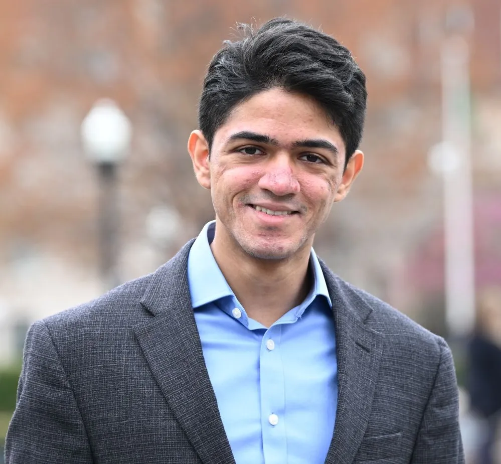 A man with dark hair in a blazer and blue button-up shirt smiles against a backdrop of a lawn and buildings.
