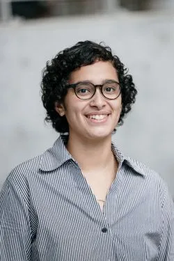 A person with short curly hair and glasses smiles warmly at the camera, wearing a striped shirt. The background is softly blurred, creating a professional tone.