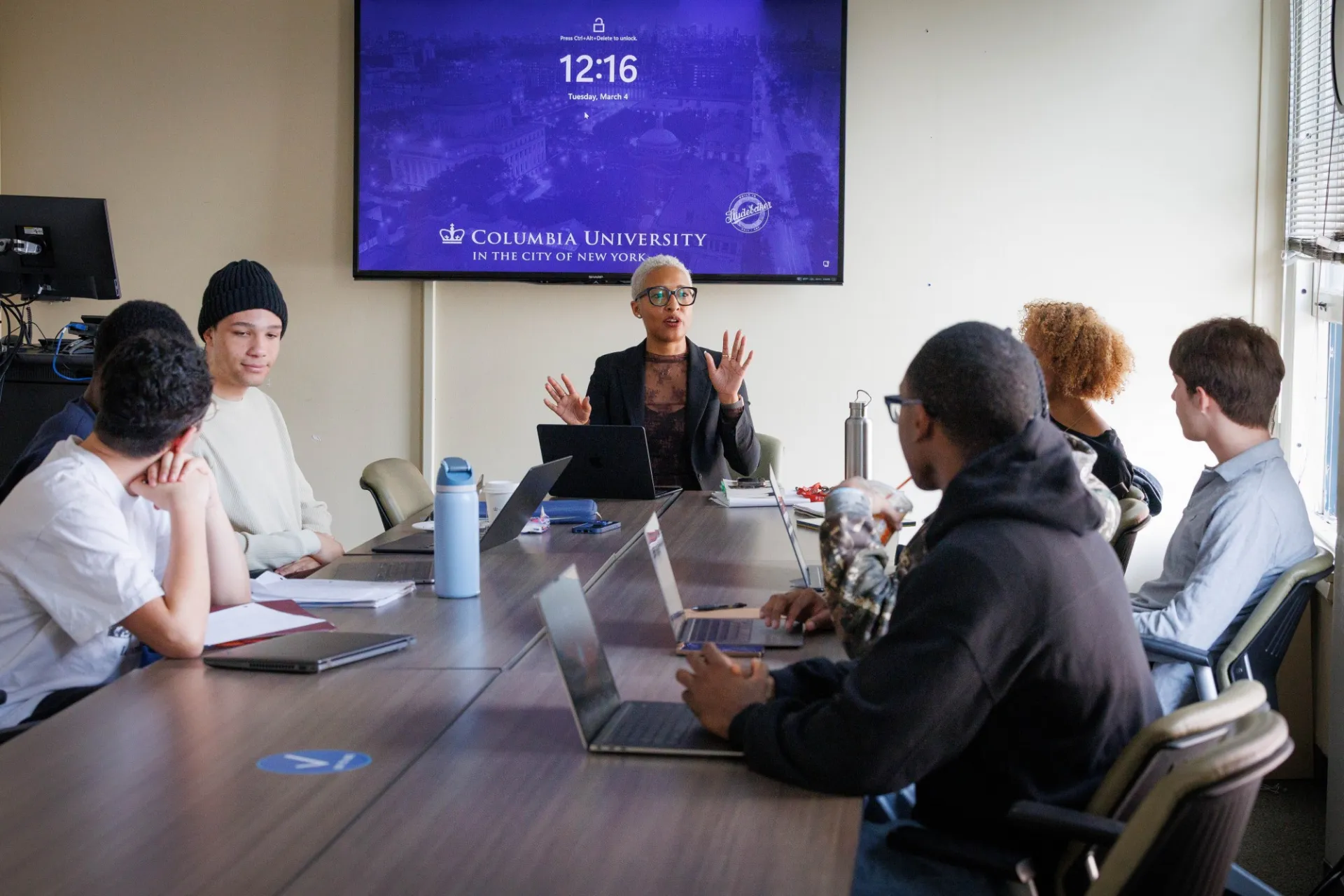 A professor speaks to her students seated around her at a table.
