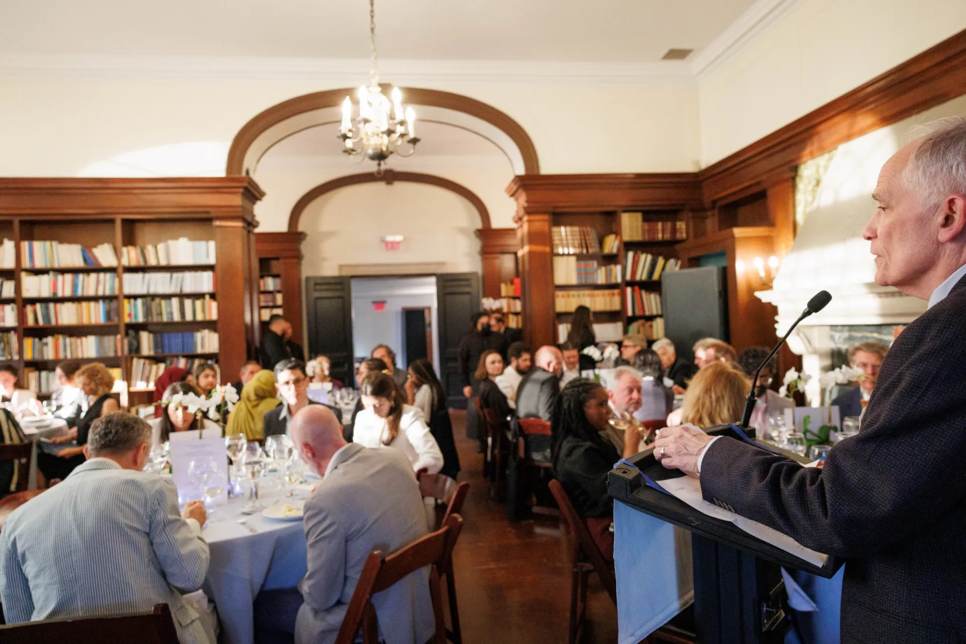 A man standing at a lectern, speaking to people seated at tables.