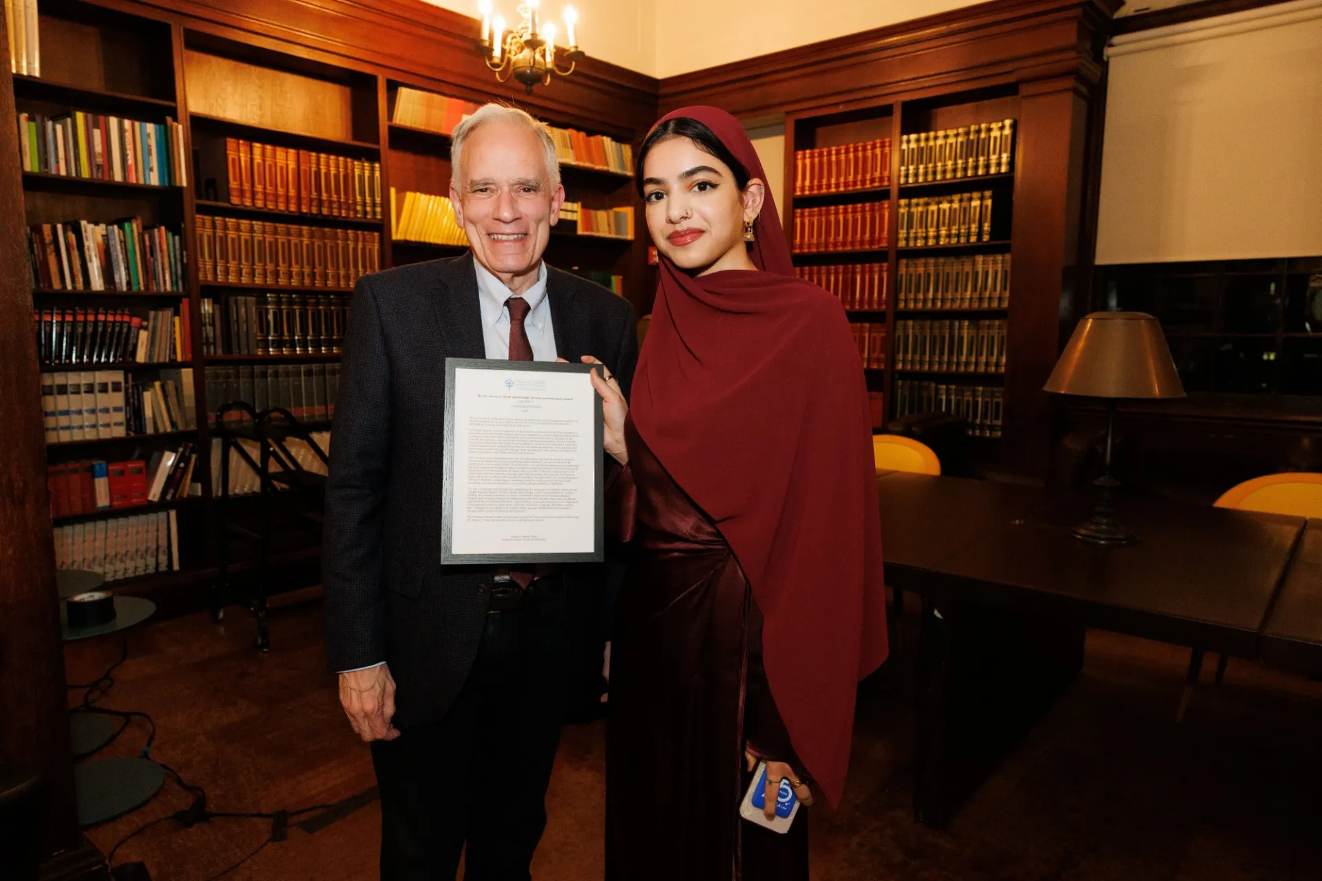 A man and a woman stand in a library holding a framed award citation.