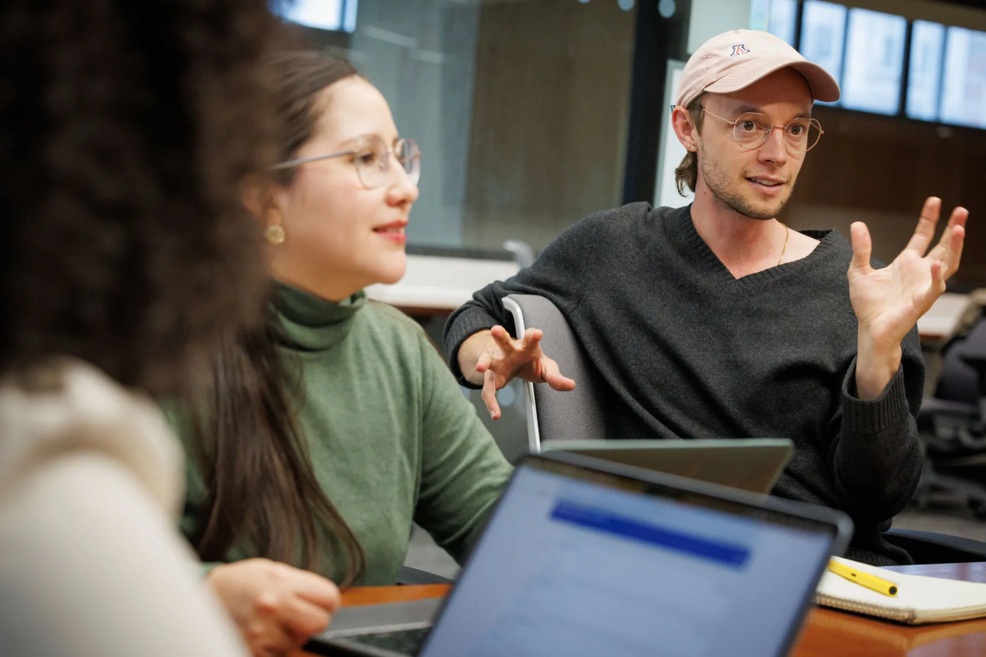 A student in a baseball cap and glasses gesticulates while speaking to classmates who are seated at a table with him, looking at their laptops.