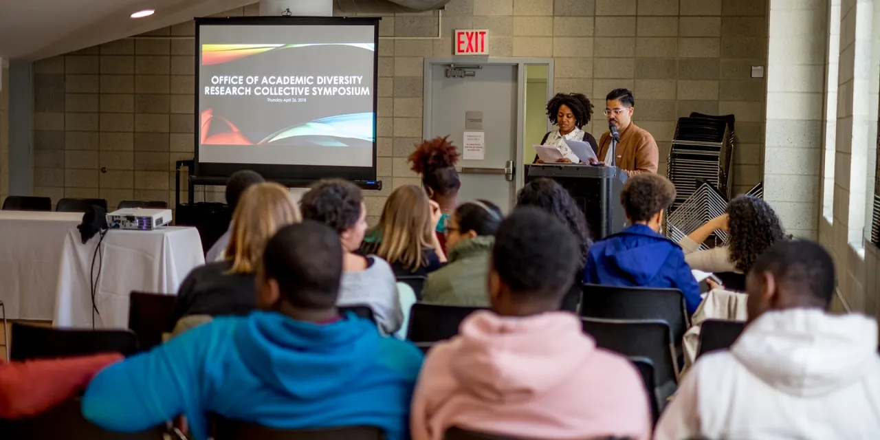 Two students at lectern next to projector screen with a presentation title slide showing, in front of an audience seated classroom style