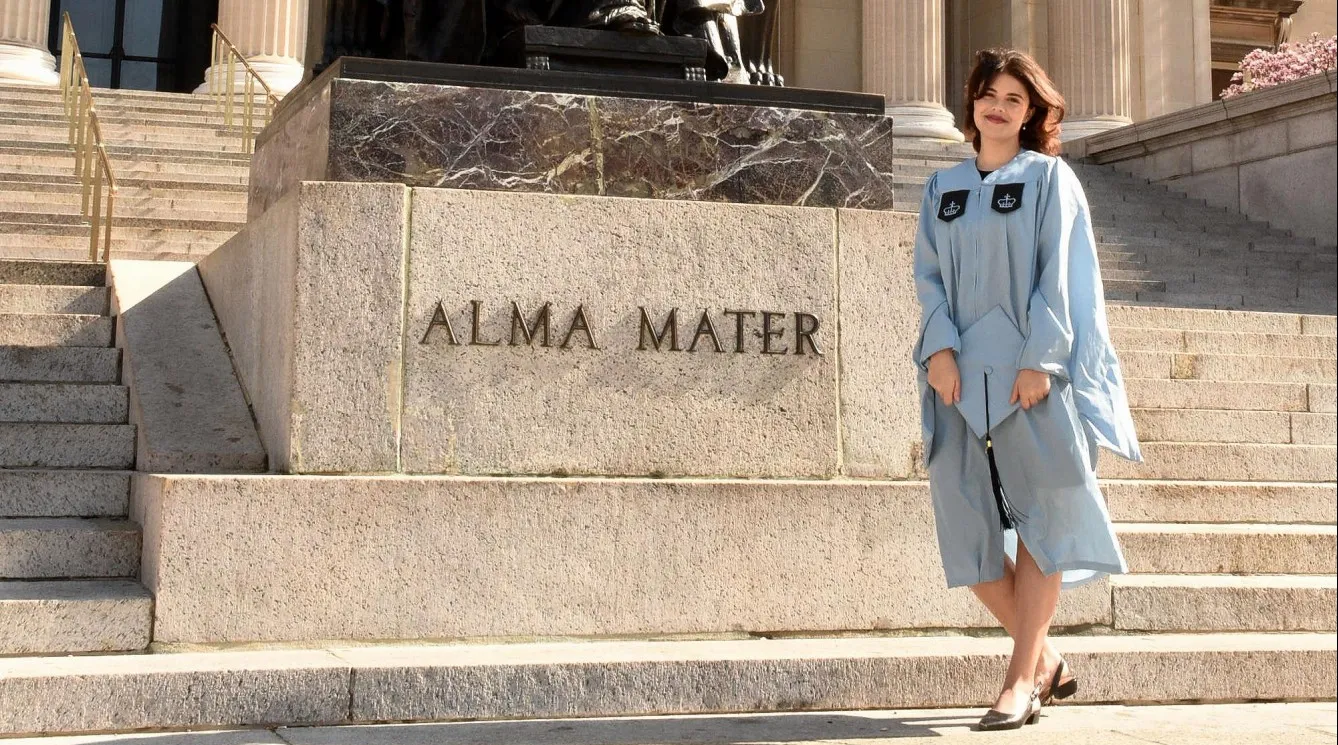 A woman in a cap and gown poses in front of the Alma Mater sculpture on the steps of Low Library. 