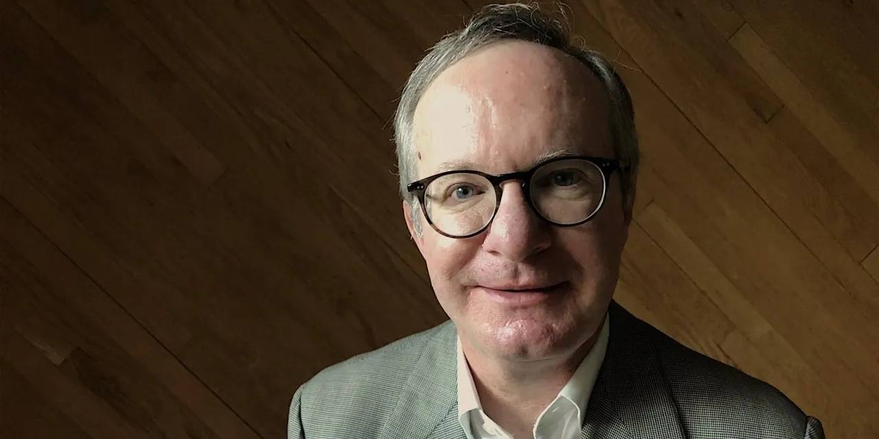 A man with glasses, gray hair, wearing a gray suit against a wood wall.