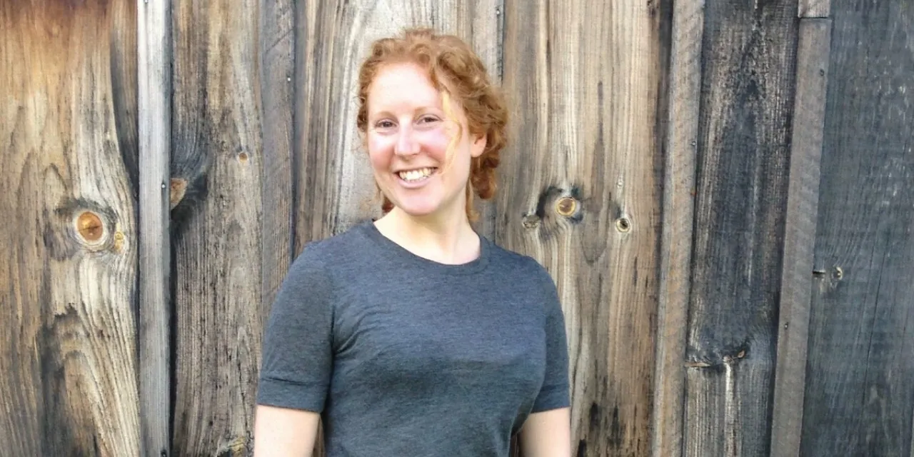 A smiling red-haired woman poses against a wooden fence.