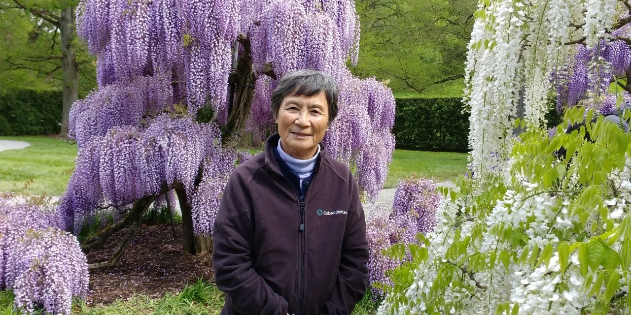 A woman in a zip-up jacket outdoors, in front of a purple wisteria tree.