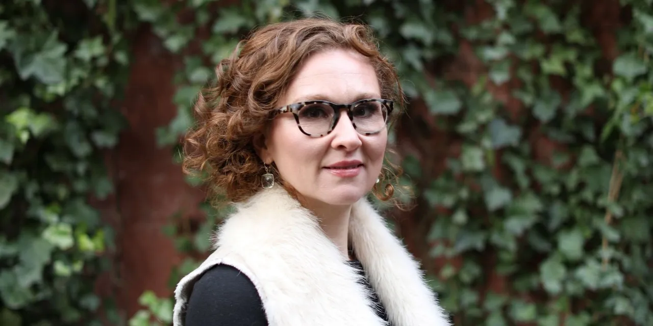 A woman with thick black glasses and curly auburn hair poses for a portrait.